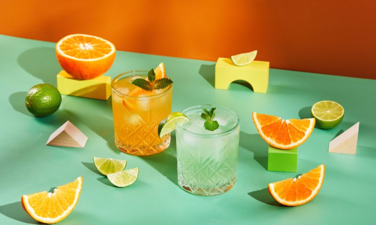 Multicolored cocktails in glasses of different shapes with orange and lime on colored backgrounds with hard shadows